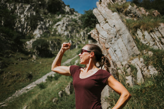 Smiling woman flexing muscles while standing against rock formation on sunny day