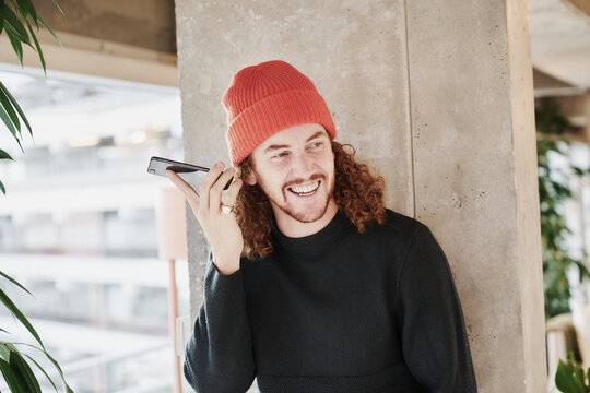 Smiling man listening audio through smart phone standing against concrete column at home