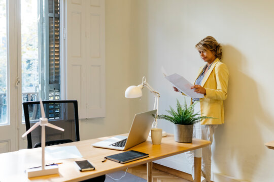 Female entrepreneur analyzing blueprint while leaning against wall in office