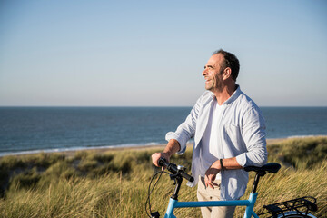 Cheerful mature man looking away while standing with bicycle at beach against clear sky