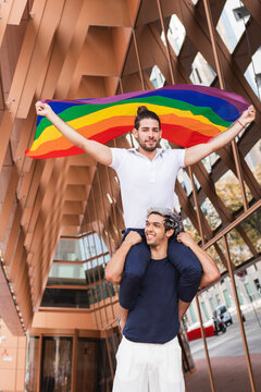 Smiling man carrying male partner with rainbow scarf on shoulders in city