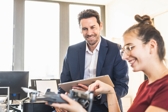 Smiling businessman with digital tablet looking at colleague holding quadcopter while sitting at office