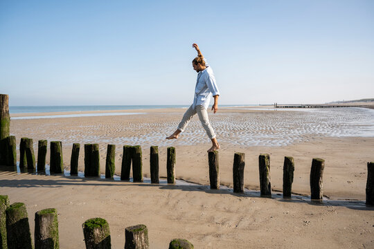 Young man balancing while walking on wooden posts at beach against clear sky - Powered by Adobe