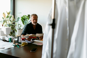 Mature male costume designer looking at fabric swatch papers while sitting at table in studio