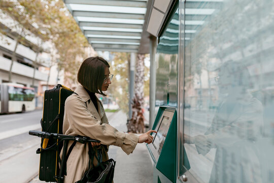 Woman with instrument case using ticket machine while standing with electric push scooter at tram station in city