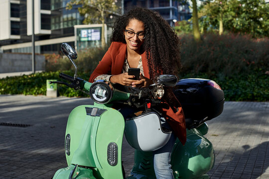 Smiling woman using smart phone while sitting on motor scooter during sunny day