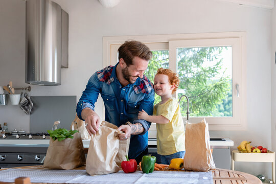 Cheerful father and son with groceries standing at dining table in kitchen