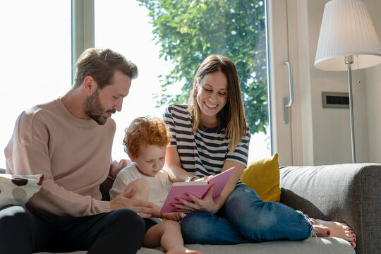 Smiling woman reading picture book while sitting by son and man on sofa in living room at home