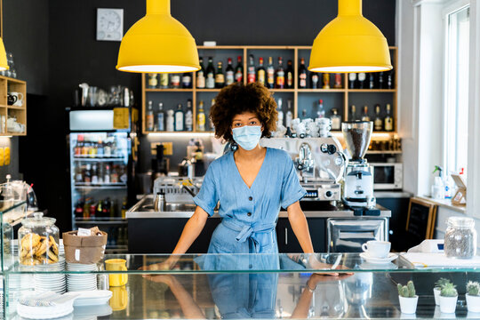 Young female owner wearing mask standing at counter in coffee shop