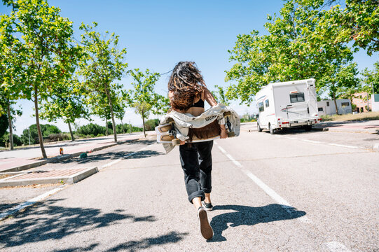 Carefree young woman carrying skateboard in the street, rear view