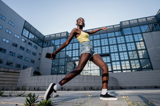 Cheerful young woman holding smart phone dancing on footpath against building in city