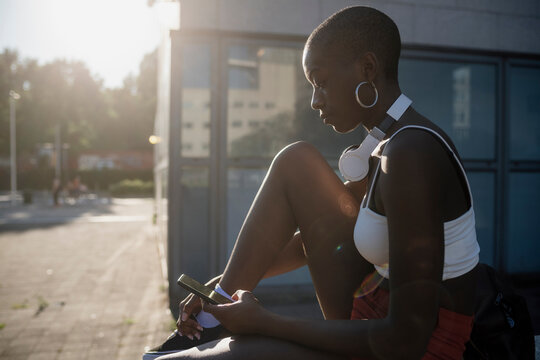 Young woman with shaved head using smart phone while sitting in city during sunny day