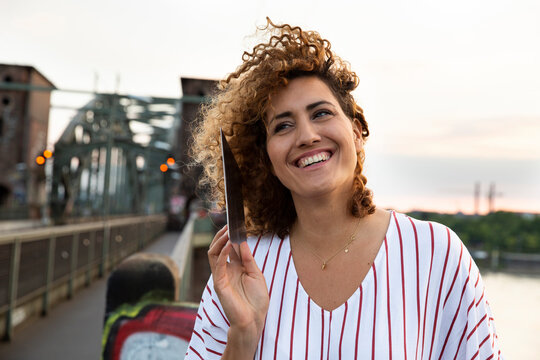 Cheerful woman with curly hair talking over digital tablet against sky in city