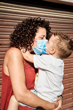 Mother carrying son with masks kissing him while standing against closed shutter