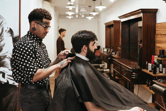 Trendy barber fixing cape on male client at salon