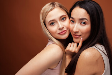 two pretty diverse girls happy posing together: blond and brunette on brown background, asian and caucasian, lifestyle people concept