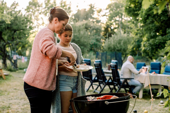Side view of mother and daughter preparing food over barbecue grill in yard
