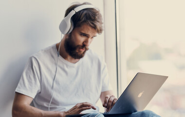 bearded man with an open laptop sits on the windowsill and headphones on his head