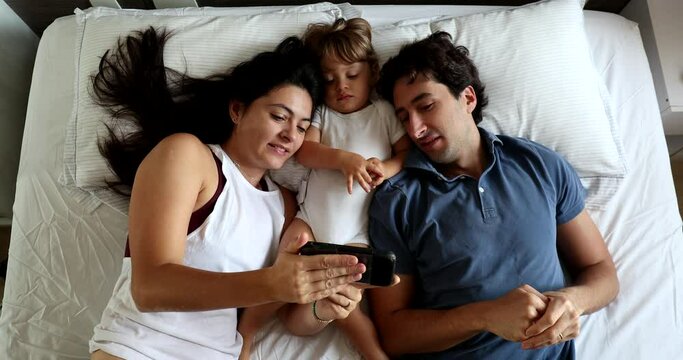 Married millennial couple watching video with infant toddler baby bou in bed