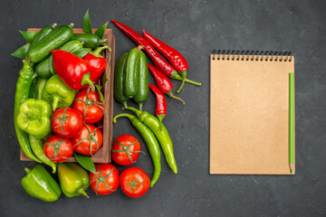 Above view of fresh vegetables inside and outside of a brown wooden basket and notebook with pen on dark background