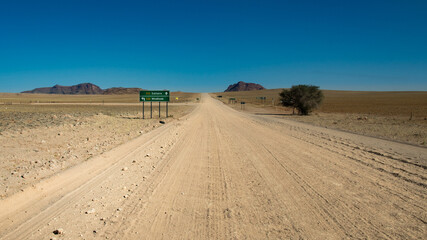 Fototapeta na wymiar road trip in Namibia - endless straight gravel road washboard in the plains of Namib Naukluft Park - intersection of two gravel roads - green road sign Solitaire-Windhoek 