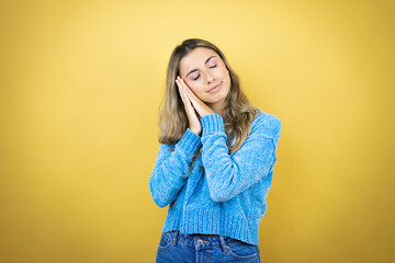 Pretty blonde woman with long hair standing over yellow background sleeping tired dreaming and posing with hands together while smiling with closed eyes.