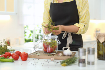Woman putting dill into pickling jar at table in kitchen, closeup