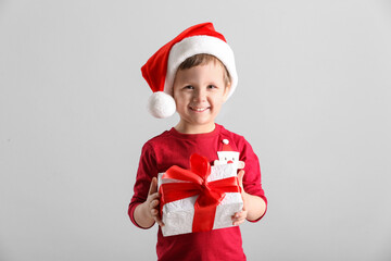 Cute little boy in Santa Claus hat holding gift box on light grey background