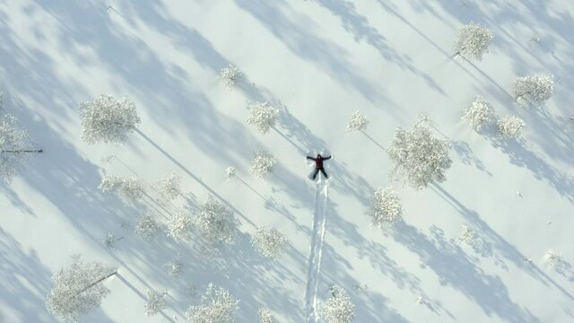 Aerial, top down, drone shot, rising over a person making snow angels, between snow covered trees, on a sunny, winter day, in Posio, Lapland, Finland