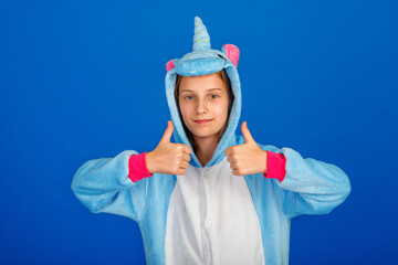 Charming girl playfully poses in the costume of the unicorn. A Studio shot of an emotional woman in kigurumi having fun on a blue background. Shows the thumbs up.