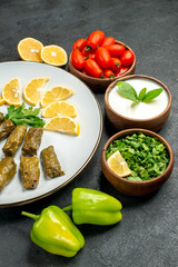 bottom view stuffed grape leaves parsley leaves and lemon half slices on plate bowls with cherry tomatoes natural yogurt green peppers slices of lemon on dark background