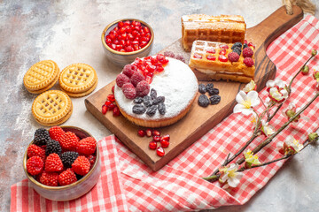 bottom view cake with berries and waffles on cutting board bowls with pomegranate raspberry candies biscuits kitchen towel on marble ground
