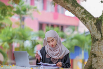 Photo of  beautiful islamic female studentwearing headscarf sitting in park and using silver laptop.