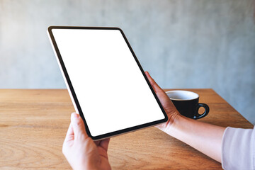 Mockup image of a woman holding digital tablet with blank white desktop screen with coffee cup on wooden table