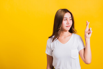 Asian Thai happy portrait beautiful cute young woman smile have superstition her holding fingers crossed for good luck and looking to camera, studio shot isolated on yellow background with copy space