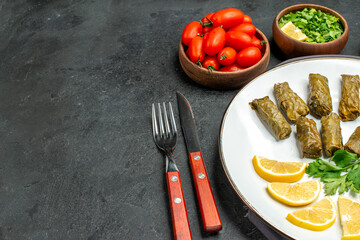 bottom half view stuffed grape leaves and lemon half slices on white oval plate bowls with cherry tomatoes parsley knife and fork and purple kitchen towel on dark background