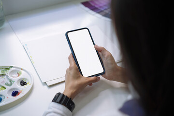 Backside view of artist woman hands using mockup white screen smart phone at desk.