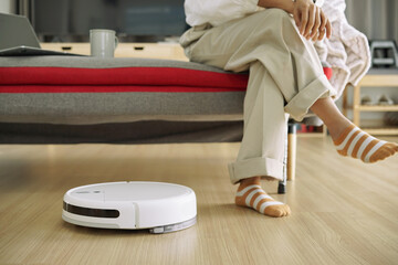 Robotic vacuum cleaner cleaning on wood floor in the living room while woman relaxing on sofa.