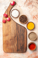 Top view of different spices lined up in a row andred radishes on brown wooden cutting board on mixed color background