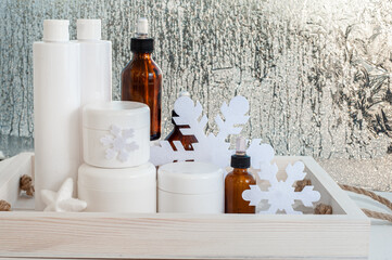 A set of winter care cosmetics against the background of the winter window with frosty patterns. White jars and vials of drips on a white table.