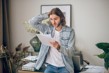 Young man in jeans shirt holding papers and looking surprised