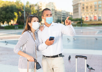 Couple of young travelers in medical masks walking around foreign city on summer day, pointing at sights. New lifestyle concept during coronavirus..