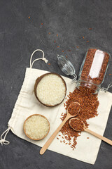 Brown rice is scattered from  glass jar onto  linen bag. Two spoons with brown rice. White rice in wooden bowls