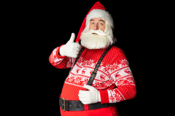 Benevolent bearded Santa Claus man in sweater and suspenders, shows well with hand with raised thumb, copying space. Isolated black background.