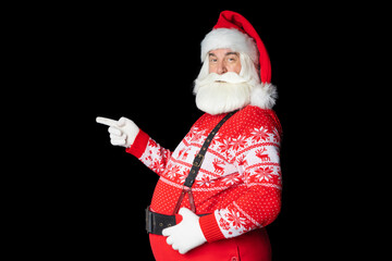 Cheerful bearded man Santa Claus in a sweater and suspenders, points a finger forward, something nearby. Isolated black background.