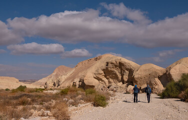 Two male friends on a hiking trail in a remote desert region. Panoramic landscape of dry wide wadi with colorful sandy hills and mountain folds . White clouds on the blue sky in a winter sunny day.