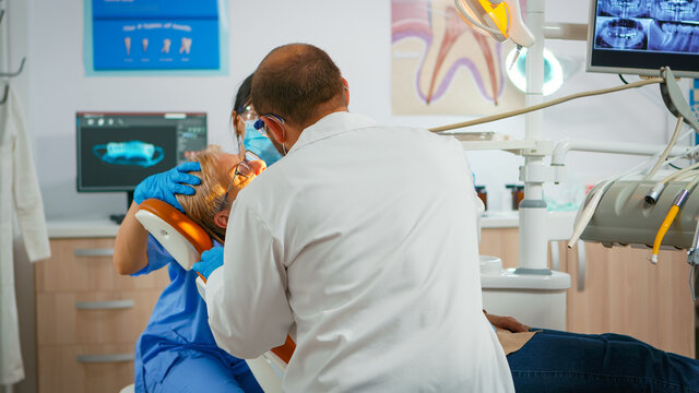 Senior woman sitting in dental clinic taking care of tooth health. Orthodontist lighting the lamp until examination, patient opening mouth lying on stomatological chair while nurse halping doctor.