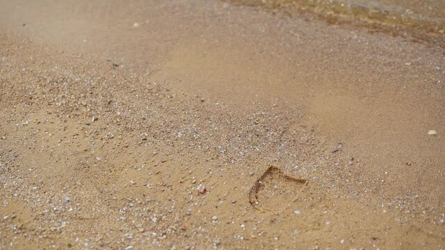 Close-up of a man feet in boots stepping on the sand near the coastline, then the man moves away and the trail remains on the ground, which is washed by the waves of the surf of the sea