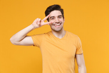 Young smiling happy satisfied unshaved caucasian handsome man 20s wears basic blank print design t-shirt showing victory sign two finger gesture near eye isolated on yellow background studio portrait.