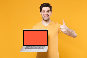 Young smiling unshaved man in basic print design t-shirt using laptop pc computer with blank screen workspace copy space mock up showing thumb up gesture isolated on yellow background studio portrait.